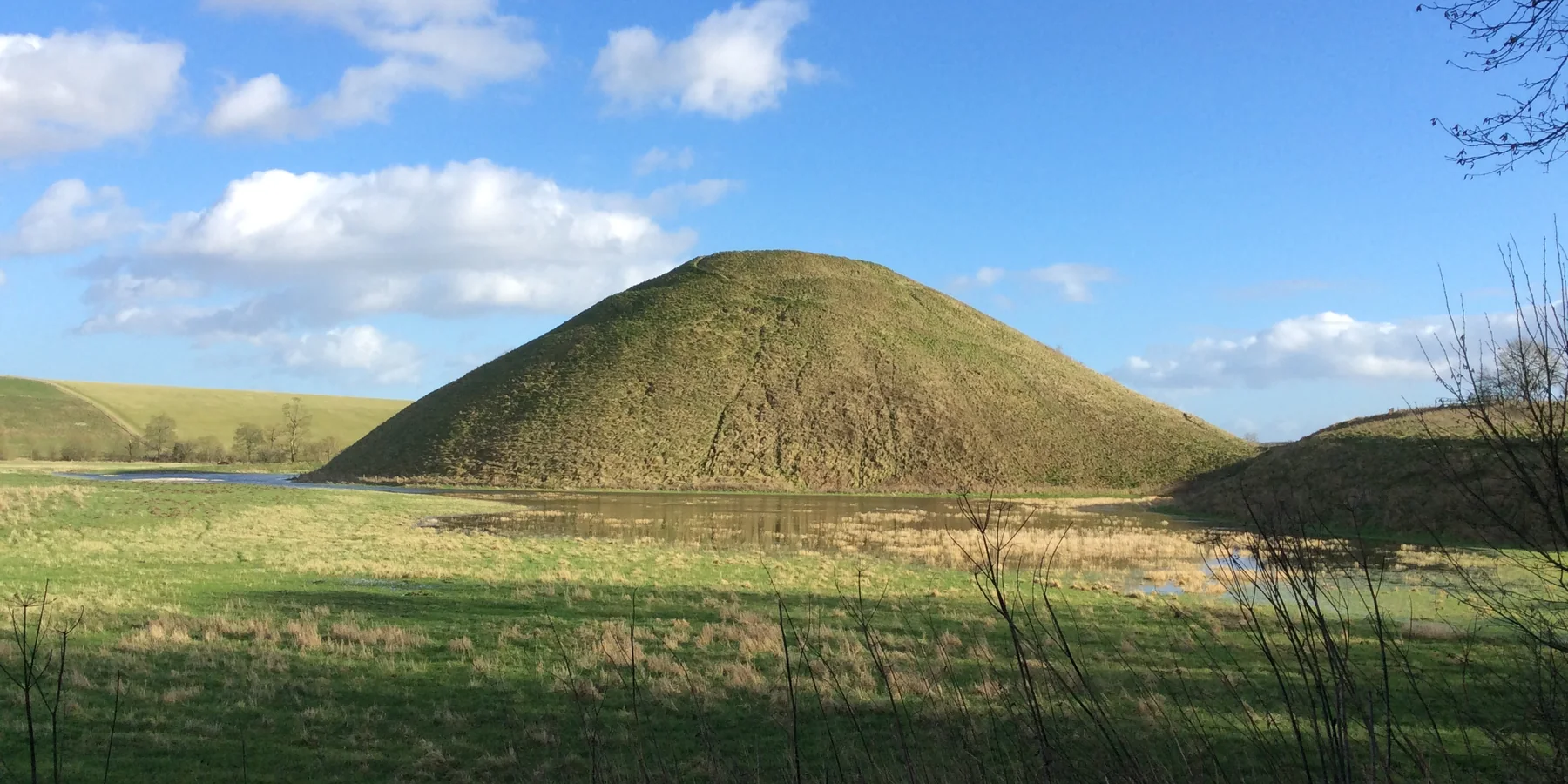 Silbury Hill, kuželovitý trávou porostlý pravěký mohylník, odrážející se v rybníce s křídovým downlandem v pozadí